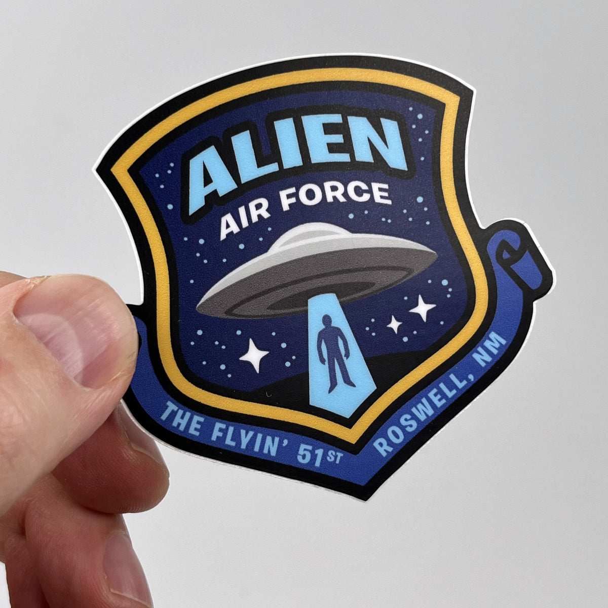 Hand holding the Alien Air Force UFO sticker, blue shield with a flying saucer