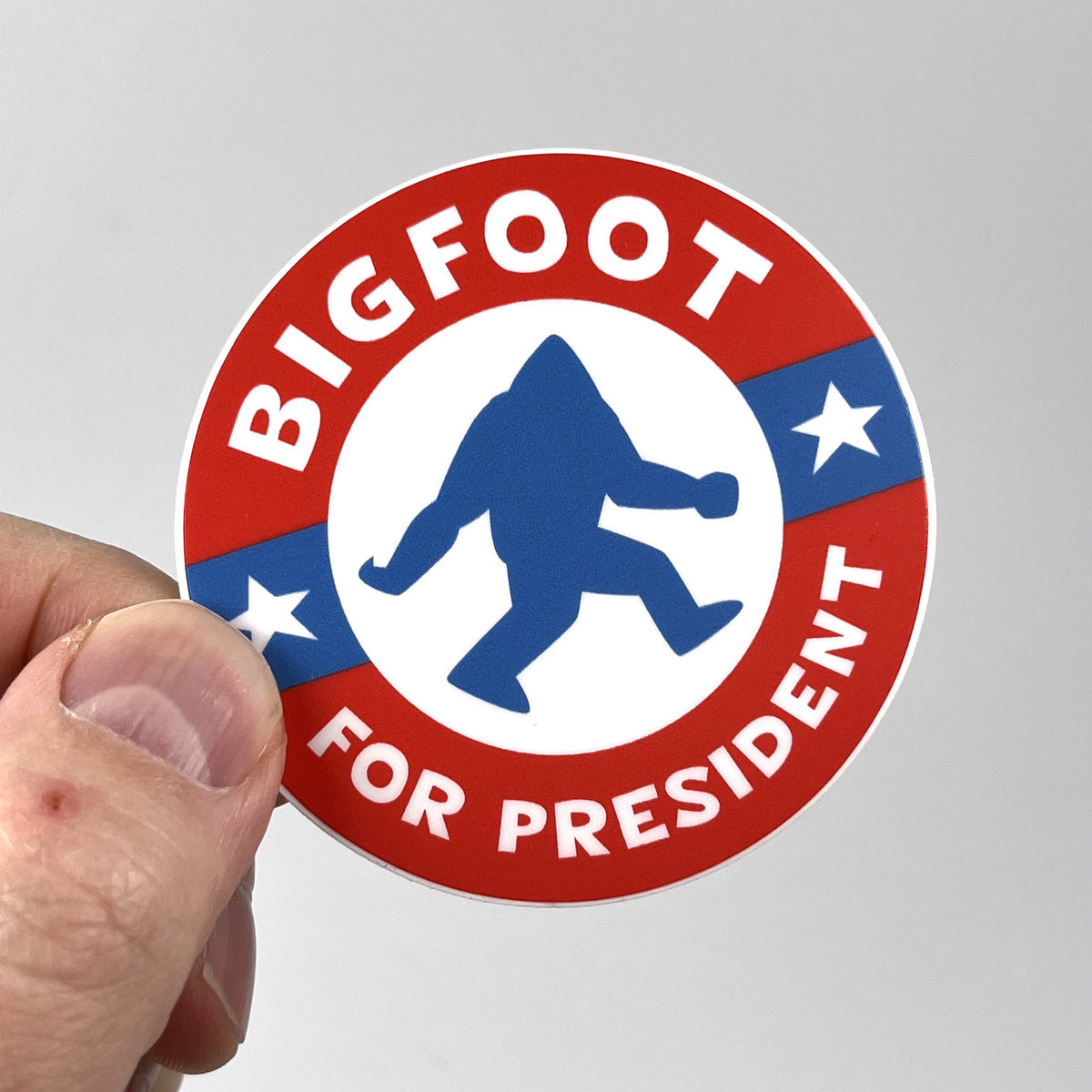 Hand holding the Bigfoot For President campaign sticker, red white and blue circle design