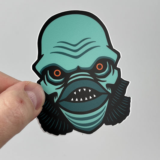 Hand holding the Lagoon Creature horror sticker, teal gill-man head