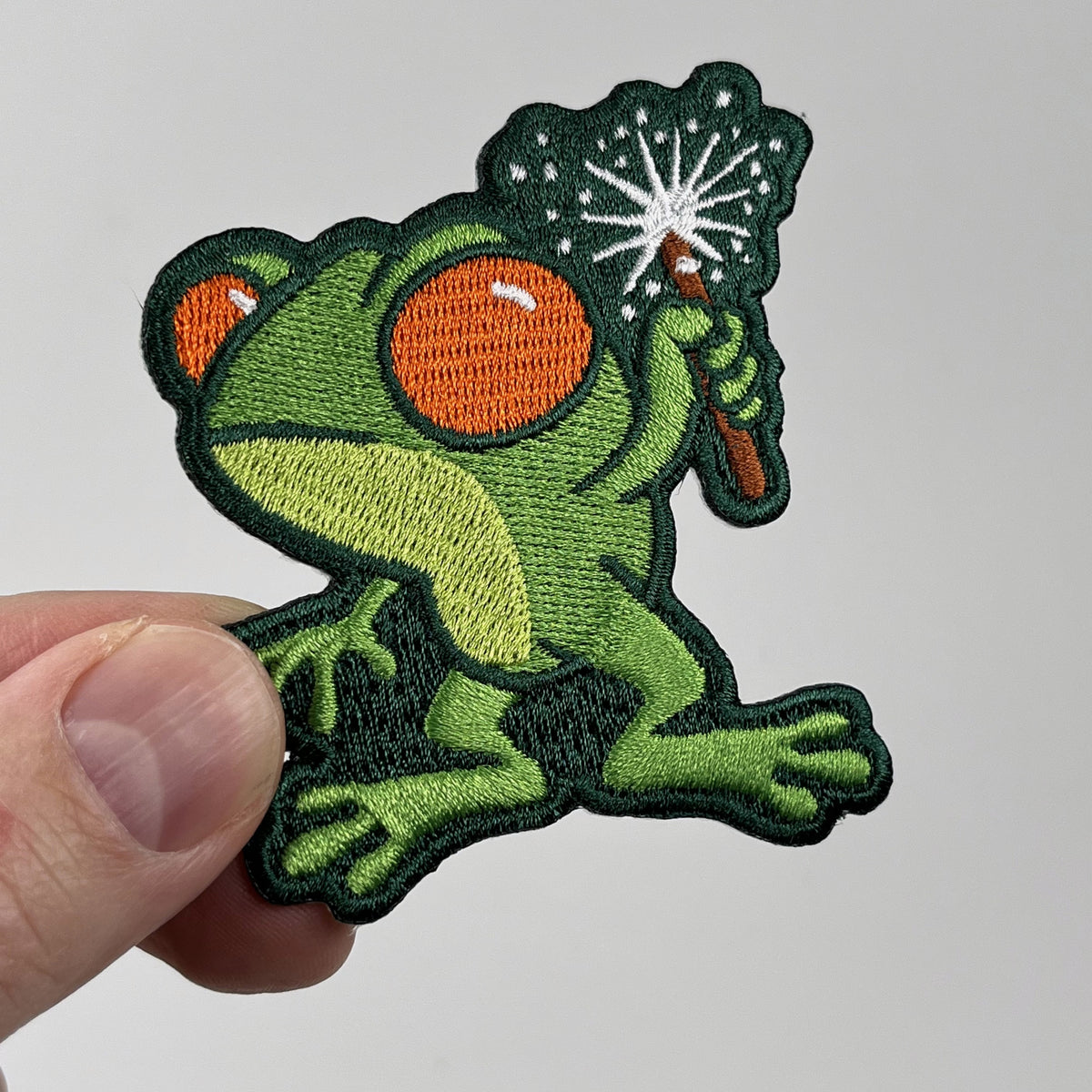 Hand holding the Loveland Frogman cute embroidered patch, green frog holding a sparkler