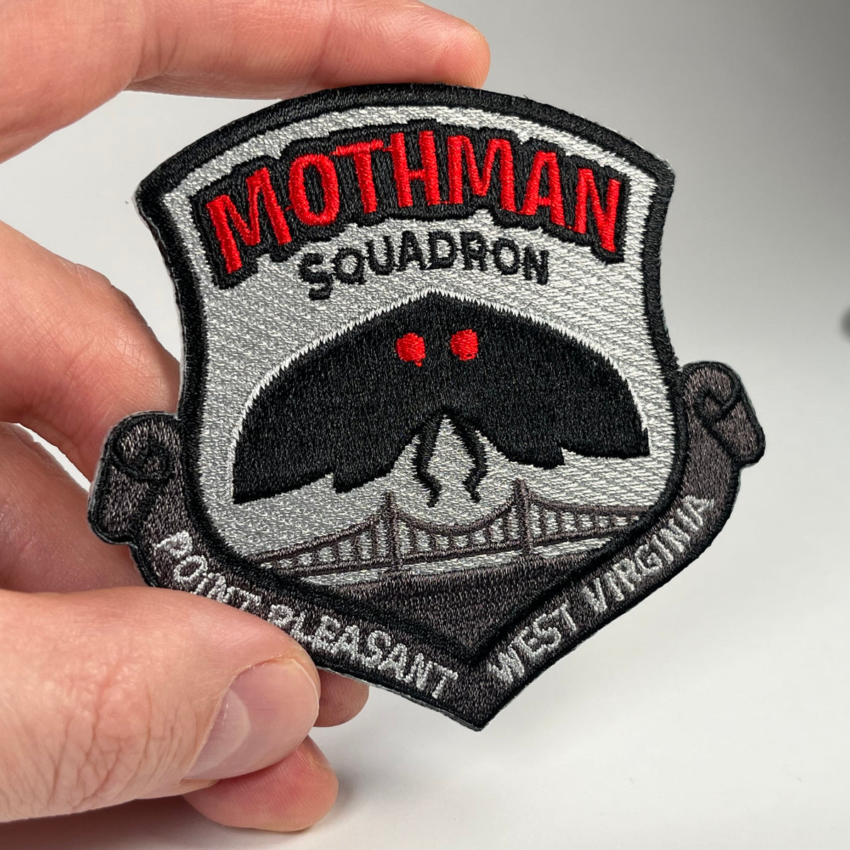 A close-up of a hand holding an embroidered 'Mothman Squadron' military badge style patch. The patch features a black Mothman cryptid with glowing red eyes over a gray suspension bridge, with 'MOTHMAN SQUADRON' in red and black text on a light gray background.