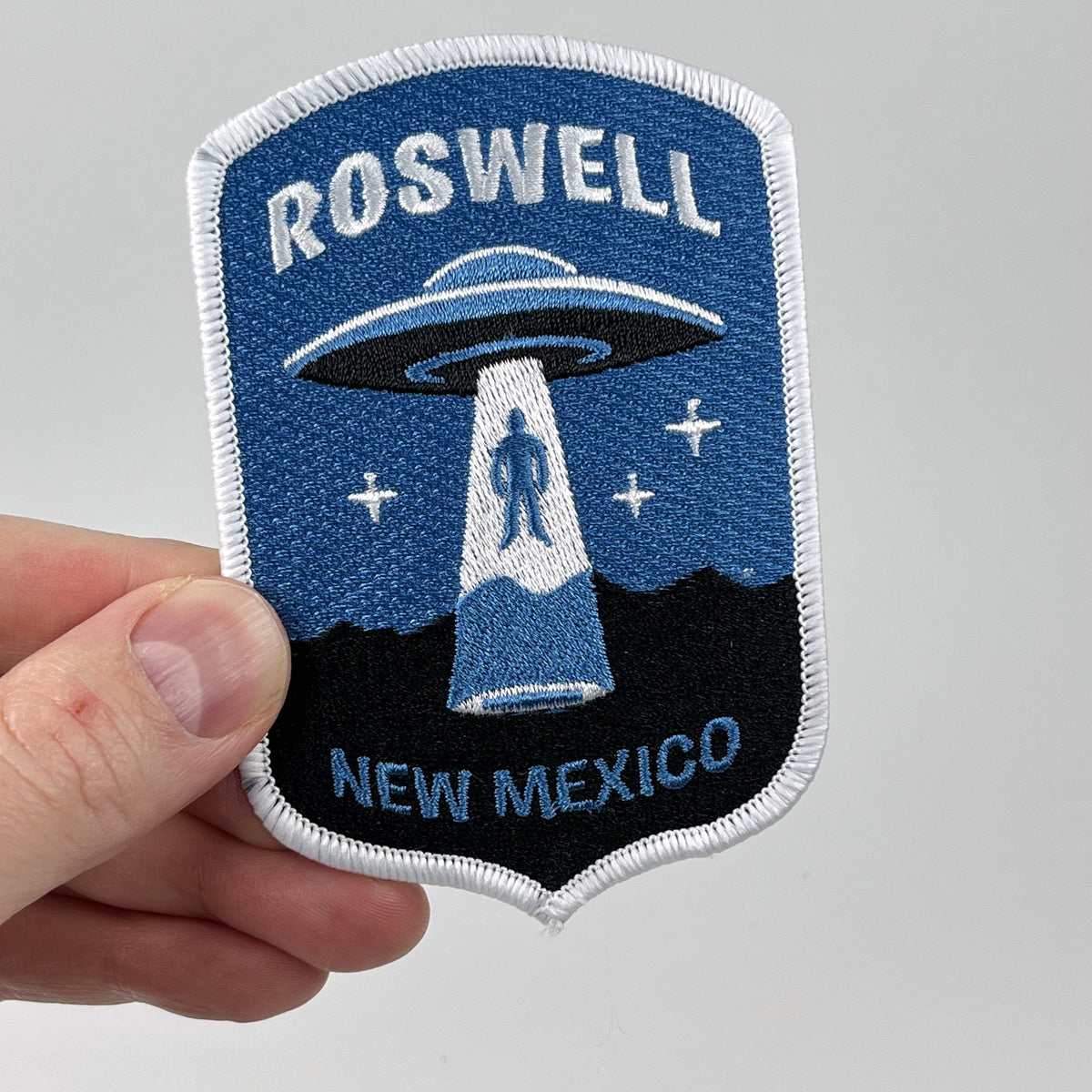 Hand holding the Roswell alien abduction embroidered patch, UFO tractor beam lifting a figure over mountains