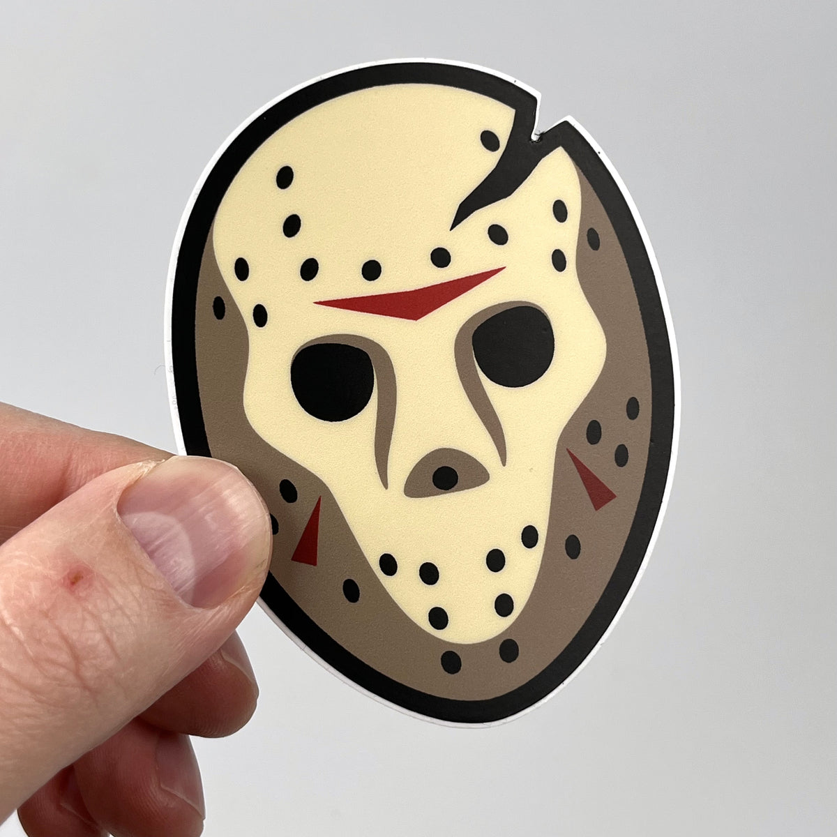 Hand holding the Voorhees horror sticker, weathered hockey mask