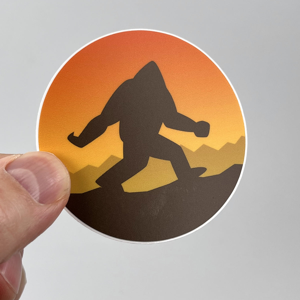Hand holding the Bigfoot Sunset cryptid sticker, Bigfoot silhouette against an orange sunset circle