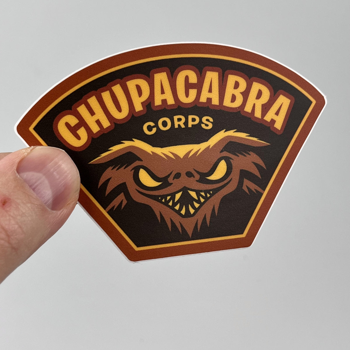 Hand holding the Chupacabra Corps cryptid sticker, brown military-style shield badge