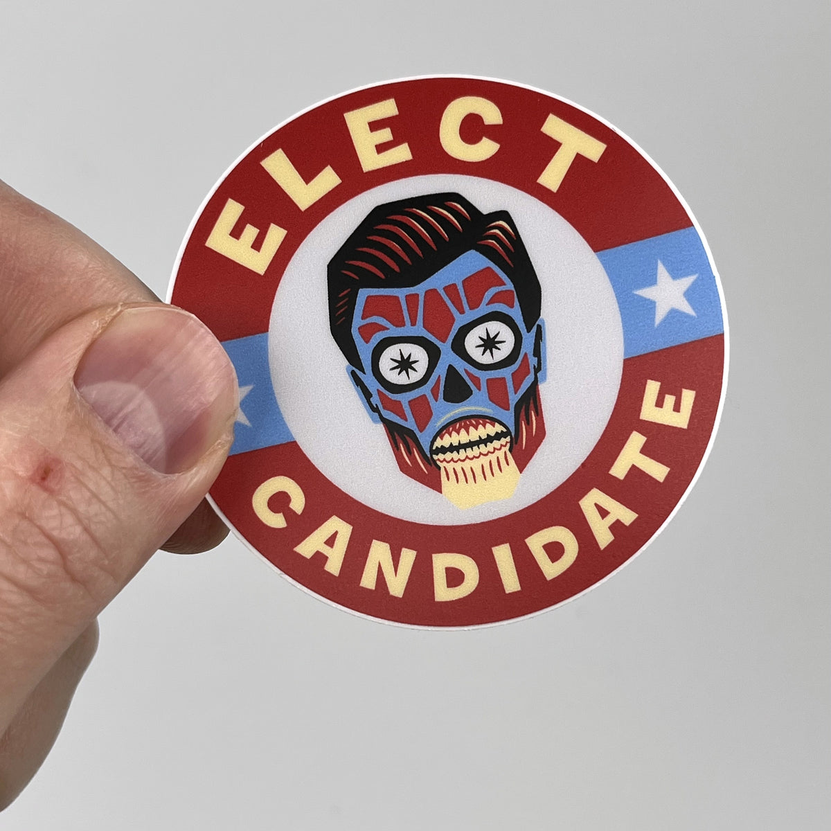 Hand holding the Elect Candidate campaign sticker, red white and blue skull design