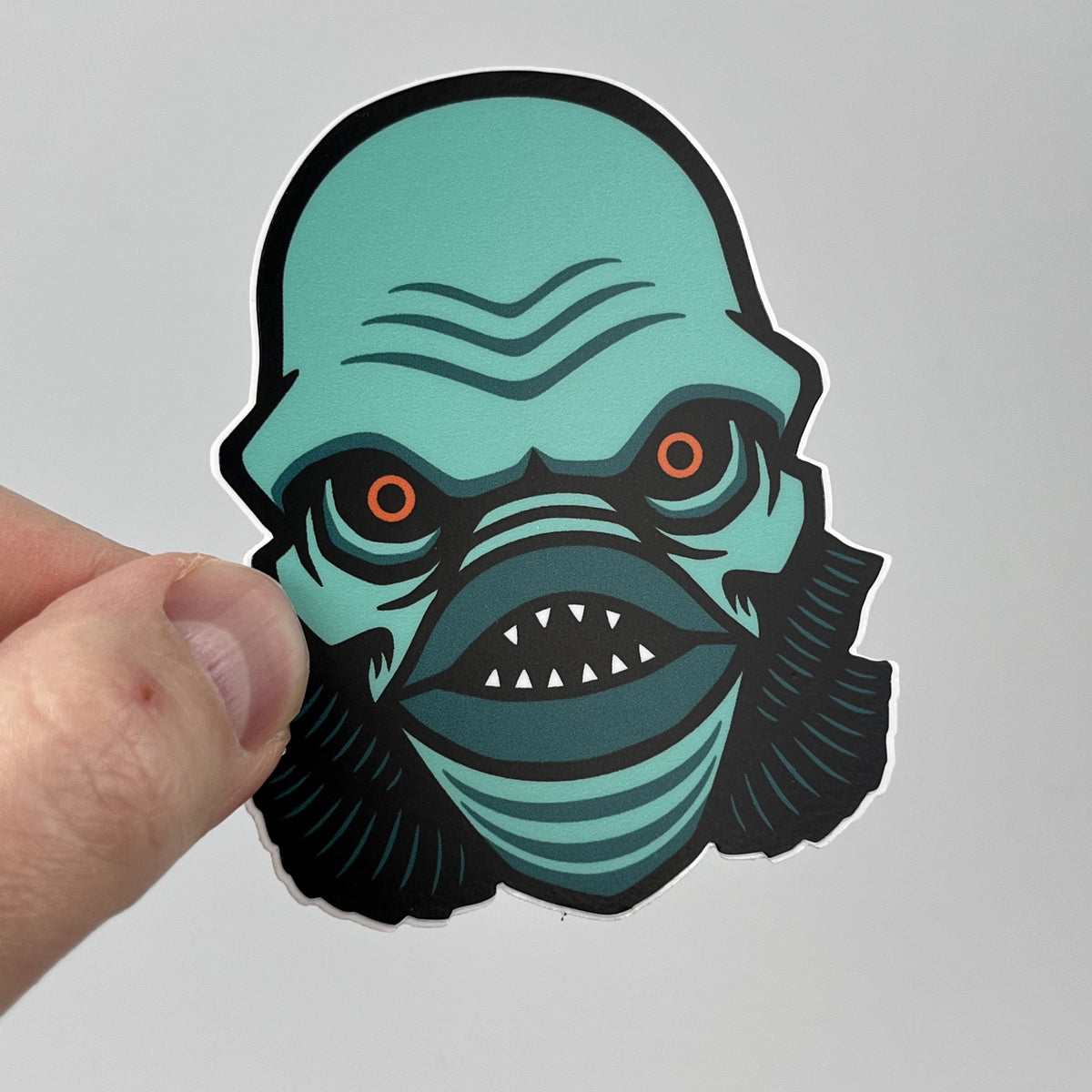 Hand holding the Lagoon Creature horror sticker, teal gill-man head