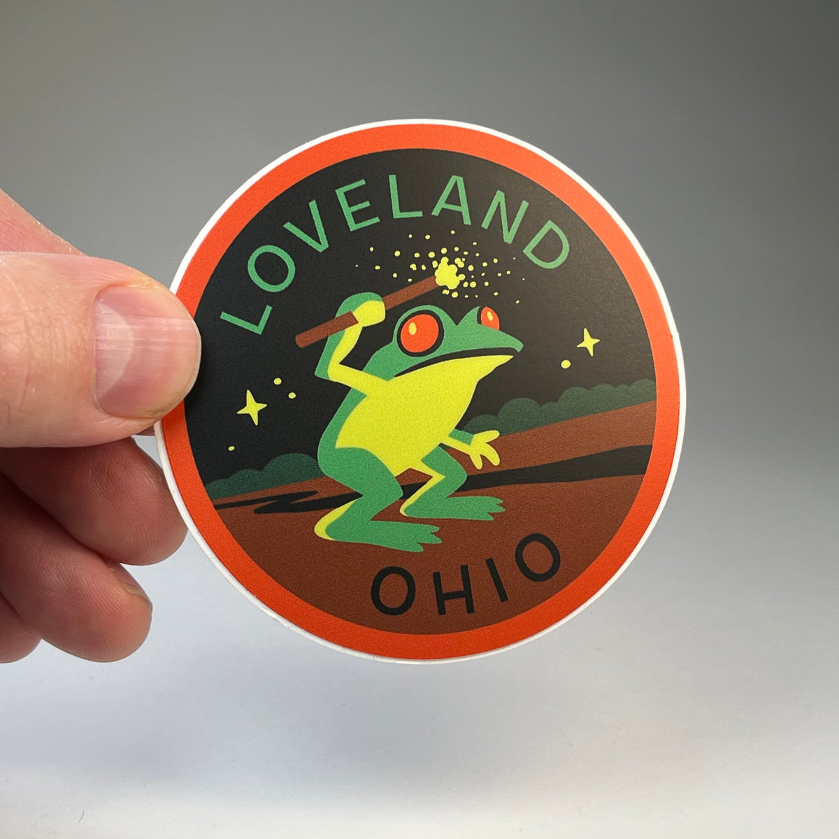 Hand holding the Loveland Ohio Frogman cryptid travel sticker
