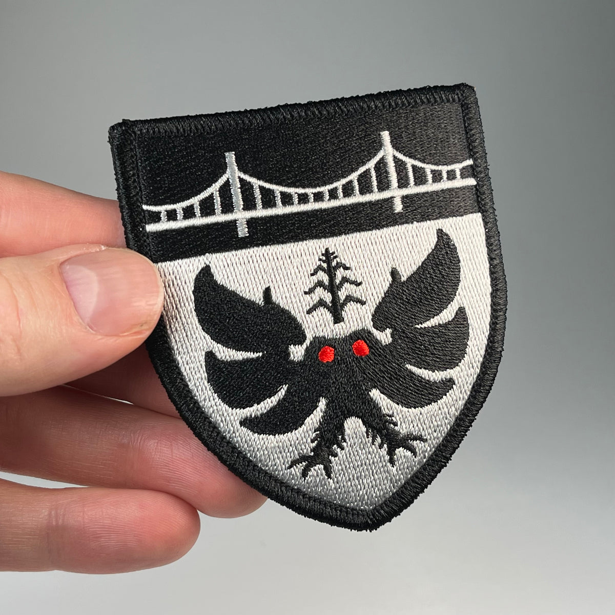 A close-up of a hand holding a shield-shaped embroidered patch featuring a black Mothman cryptid with red eyes, and a white bridge on a black and white background, against a neutral silver backdrop.