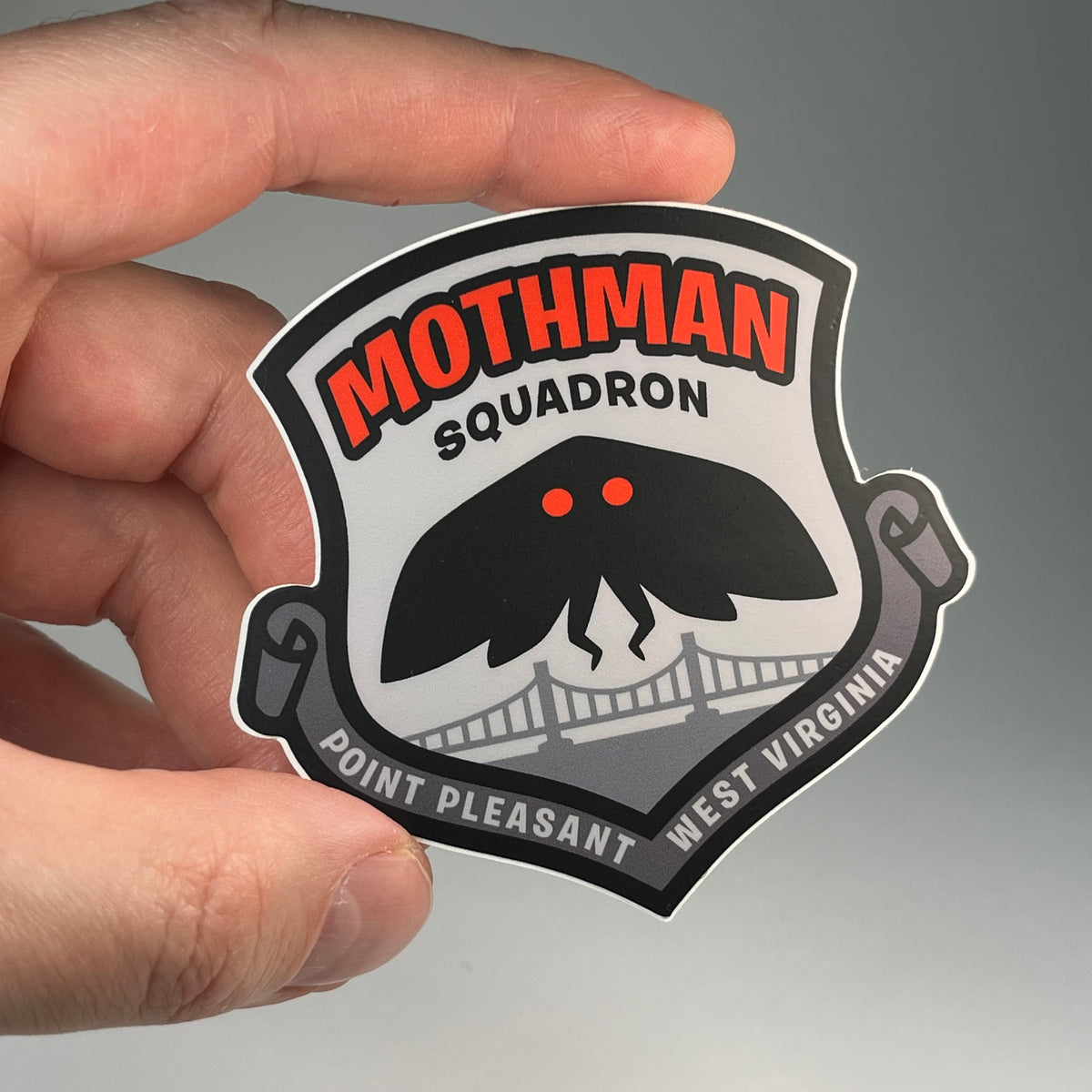 A hand holding a die-cut sticker featuring the 'Mothman Squadron' emblem from West Virginia. The sticker has a white background with a black border, red lettering, and a black Mothman cryptid graphic with red eyes above a bridge, with 'Point Pleasant West Virginia' written along the bottom.