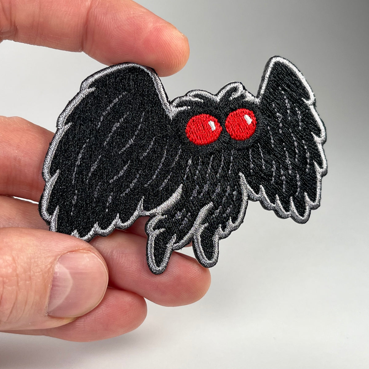 A hand holding a black embroidered Mothman cryptid patch with bright red eyes and a silver-gray stitched outline, shown against a plain light gray background.