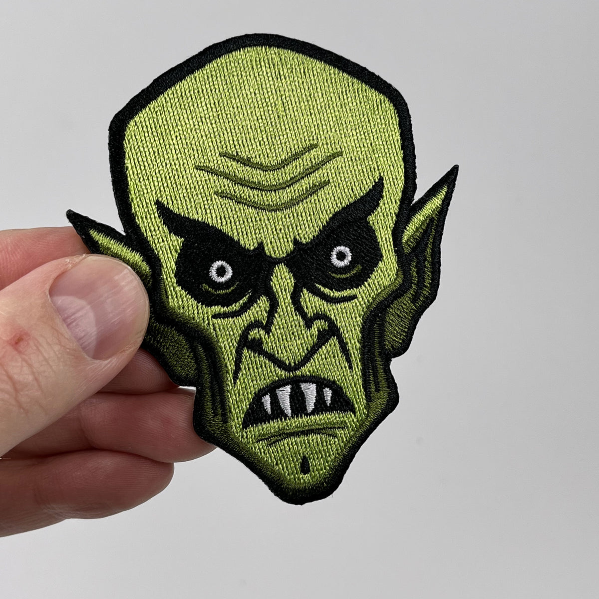 Hand holding the Orlok Nosferatu vampire embroidered patch, green bald vampire head with pointed ears