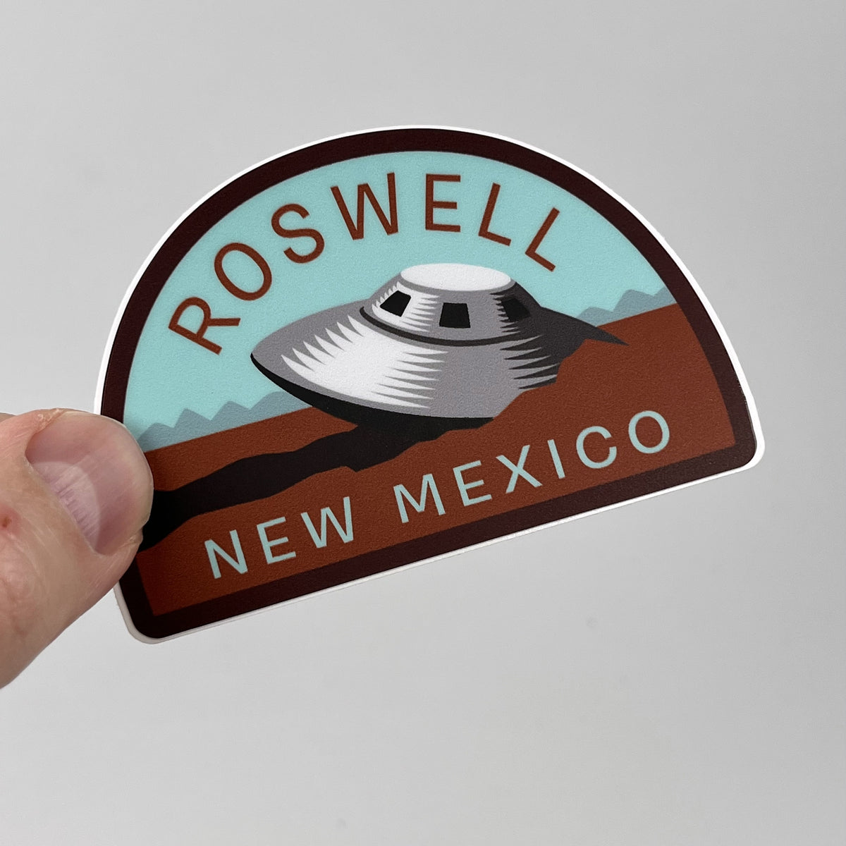 Hand holding the Roswell New Mexico UFO travel sticker, flying saucer over desert mountains