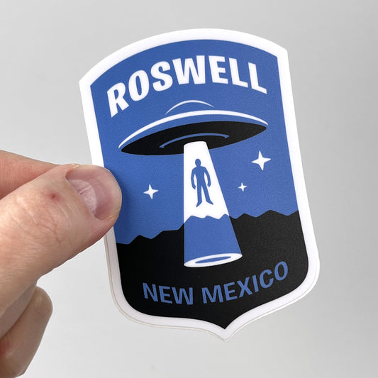 Hand holding the Roswell alien abduction UFO sticker, blue shield with a tractor beam lifting a figure
