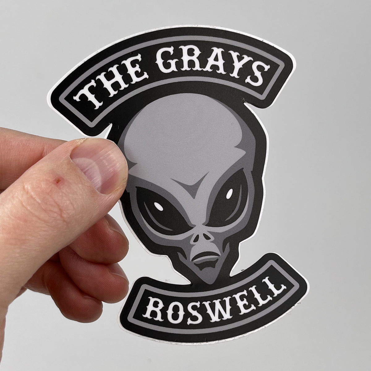 Hand holding the Grays motorcycle club alien sticker, gray alien head with MC-style rocker banners