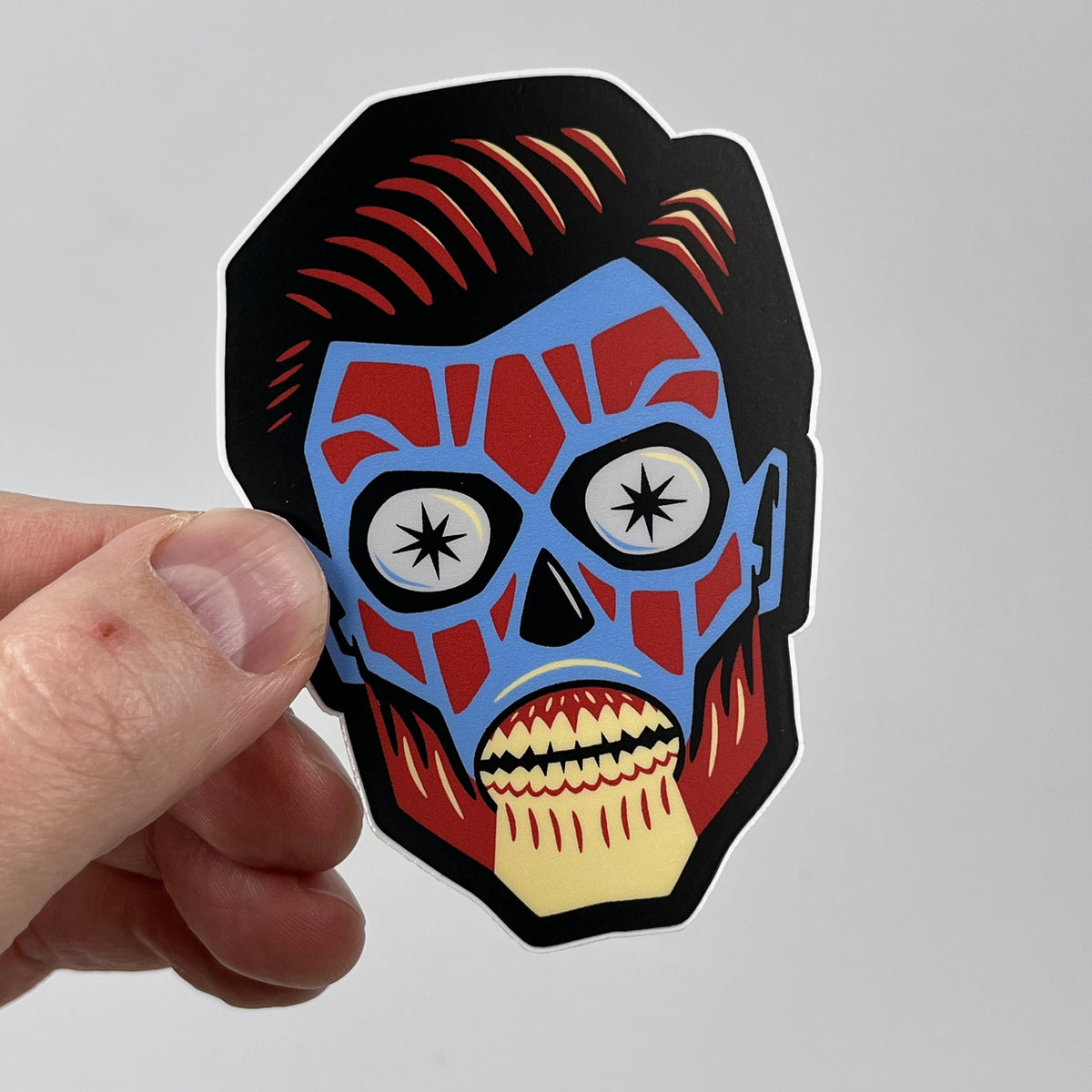 Hand holding the They Live alien head sticker, blue and red movie monster face