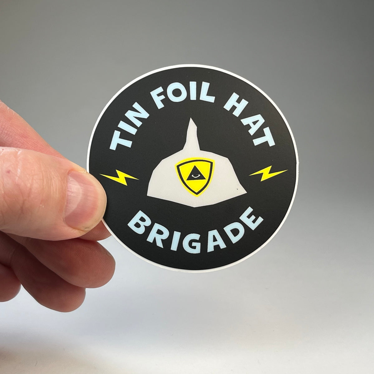 Hand holding the Tin Foil Hat Brigade circle sticker